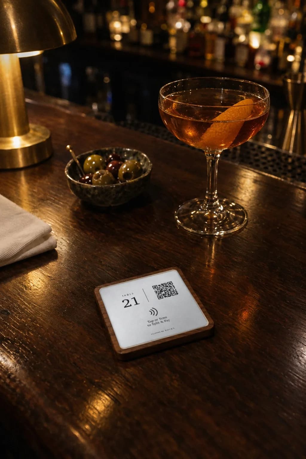 A RHYKA plaque for table 21 lying on a dark wood bar next to a stirred cocktail