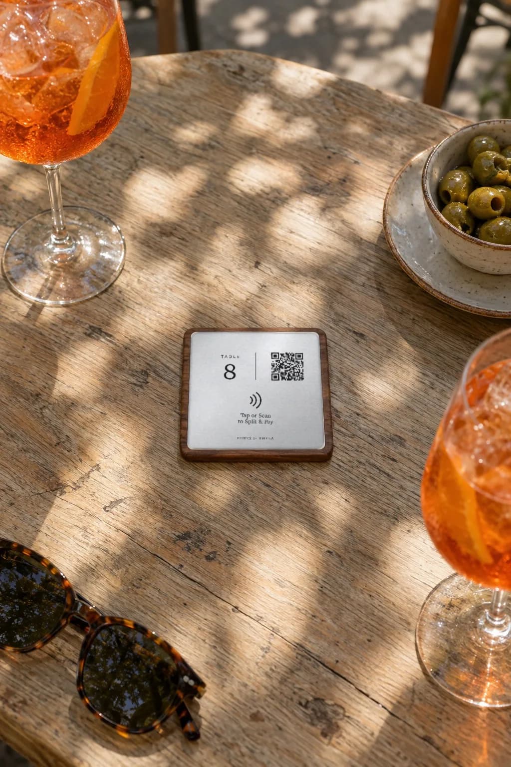 A RHYKA plaque for table 8 lying in the middle of a sunny patio table, spritzes and olives around it